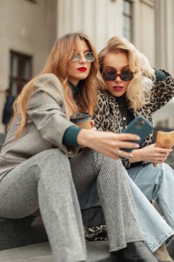 Moda kıyafetleri içinde, klasik güneş gözlüklü iki güzel moda genç kız kardeş basamaklarda oturur, telefonda selfie çeker ve kahve içerler. Seyahat ve şehir tatili kavramı