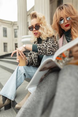 İki moda güzel sarışın ve süslü elbiseli kızıl saçlı bir kız şehirde oturmuş, kahve içiyor ve dergi okuyorlar. Moda, Güzellik ve Tarz
