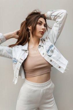 Modaya uygun kot elbiseli, ceketli, bluzlu ve pantolonlu güzel bir kız model sokakta gri bir duvarın yanında duruyor.