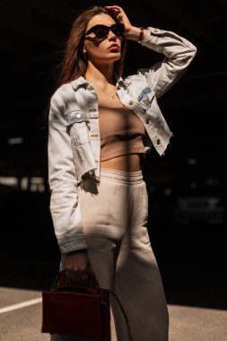 Moda modası geçmiş, modaya uygun, kot ceket, pantolon ve deri çantalı güneş gözlüklü beyaz kız modeli gün ışığında sokakta yürüyor.
