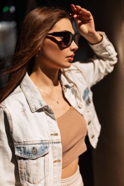 Güneş gözlüklü, kot ceketli ve güneş ışığında sokakta duran güzel bir kadın modelinin güneşli portresi.