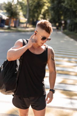 Havalı yakışıklı hippi, güneş gözlüklü, saç stili siyah moda yaz kıyafetleri giyen, sırt çantalı, sokakta yürüyen ve kablosuz kulaklıklı müzik dinleyen biri.