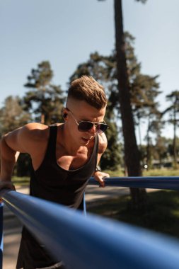 Havalı yakışıklı sporcu güneş gözlüklü siyah elbiseli vücut geliştirici açık havada yatay barda şınav çekiyor.