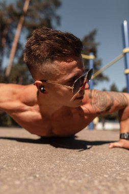 Kaslı vücutlu, havalı, güçlü, güneş gözlüğü takan, kablosuz kulaklık takan, spor yapan ve asfaltta şınav çeken bir sporcu.