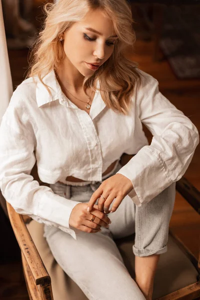 Moda kıyafetleri içinde güzel sarışın manken odasındaki tahta koltukta oturup dinleniyor.