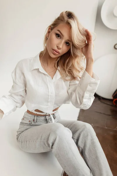 工作室里坐着一位性感漂亮的金发女模特儿 穿着时髦的白色衬衫 牛仔裤 直着头发 看着镜头 — 图库照片
