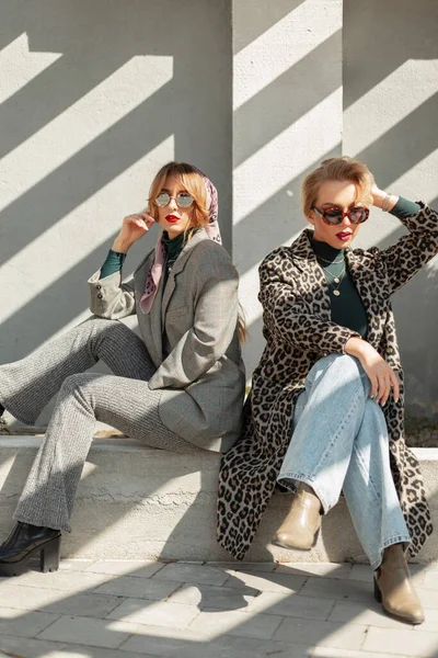 两个穿着时髦衣服 戴着太阳镜的年轻貌美女子坐在混凝土墙边的太阳光下 妇女的复古都市风格和时尚 — 图库照片