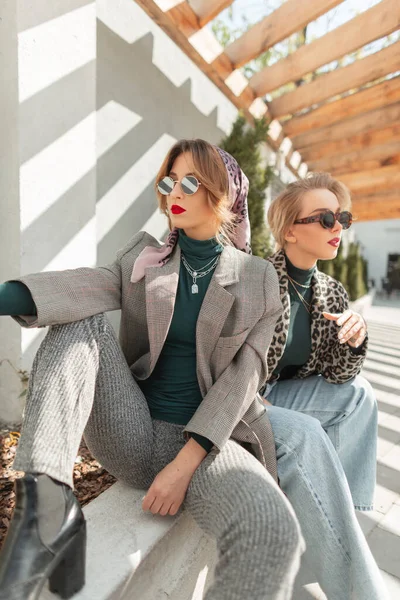 流行のグレーのスーツにスタイリッシュなサングラスを持つかなりファッション若い女性は セーターや靴は通りにスタイリッシュなガールフレンドと座っています 都会の女性の秋の服 — ストック写真