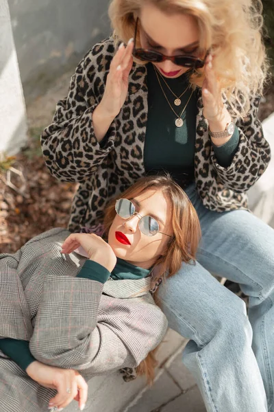 两个时髦漂亮的嬉皮士女人 戴着老式太阳镜 穿着时髦的春装 外套和牛仔裤坐在外面 女人的时尚 美丽和时尚 女朋友们 — 图库照片
