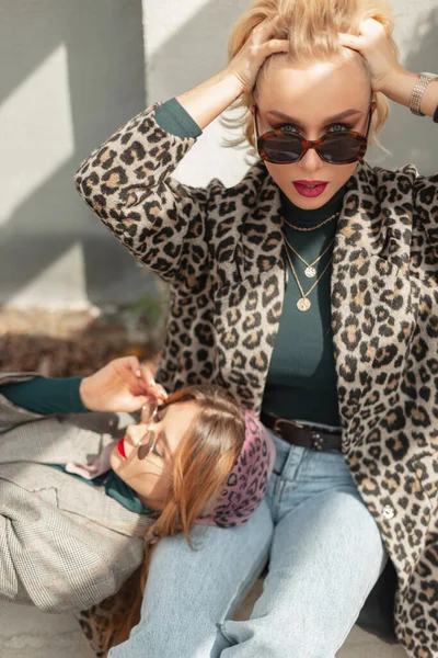 Güneş gözlüklü, leopar desenli ve kot pantolonlu güzel bir manken kadın hipster kız arkadaşlarıyla oturuyor ve sokaktaki kameraya bakıyor.