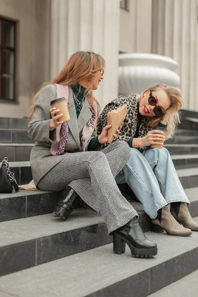 Fashion funny happiness women in trendy stylish clothes with shoes sits on steps and drinks coffee in the city. Travels and happy time
