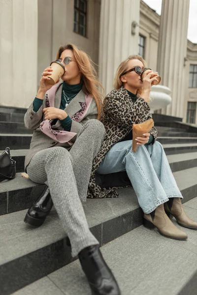 Two fashion happy urban girls hipster in stylish trendy autumn clothes with shoes travels and sits on the steps in the city, drinks coffee in cups