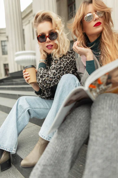 Two fashionable girls in fancy clothes sitting on the steps, drinking coffee and reading a magazine in the city