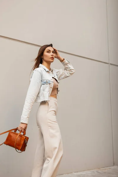 おしゃれな革のハンドバッグと流行のジーンズの服のファッションの美しさ若い女性は灰色の建物の近くの街を歩く — ストック写真