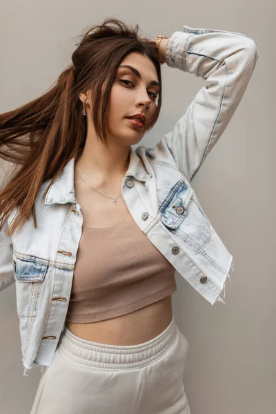 Happy fashion pretty girl in jeans jacket with top and pants stands near a gray background on the street. Urban female denim style clothes