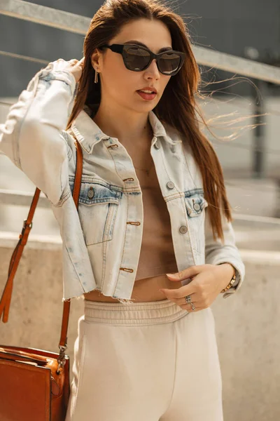 Havalı moda hippi kız şık güneş gözlüklü şık kot ceketli, üst ve pantolonlu şehirde çanta taşıyan.