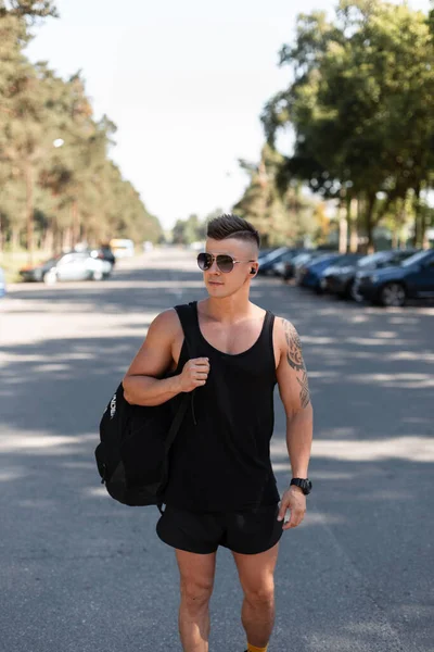 身穿黑色夏装 背着背包 留着发型 戴着太阳镜的时髦年轻帅哥走在一个有停车场的公园里 — 图库照片