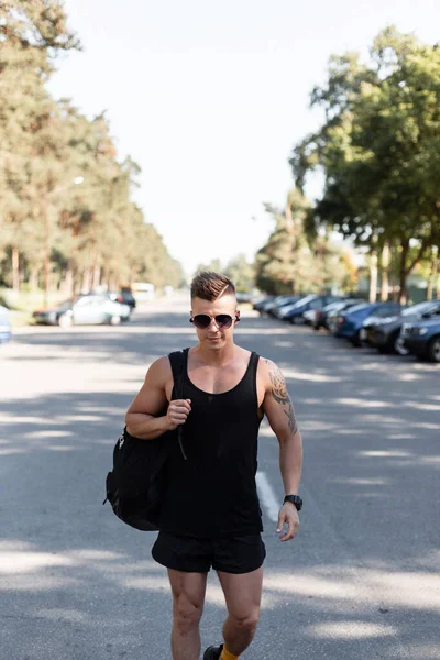 sporty handsome man model with a hairstyle and vintage sunglasses in fashion black clothes with a backpack walks on the street and listens to music