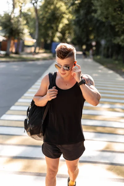 handsome fashionable man with a haircut and sunglasses in a summer fashionable black outfit  with a backpack walks and listens to music on headphones outdoors