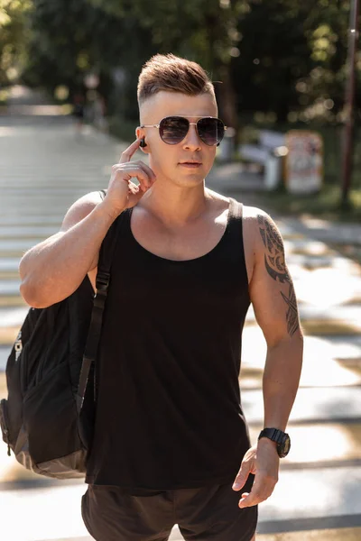 Fashion man model with hairstyle and sunglasses in black stylish summer clothes with backpack listening to music and walks on the street. Male urban style outfit