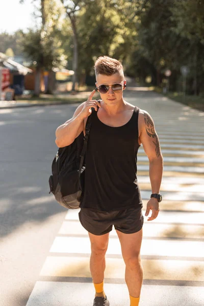 Sports handsome hipster man with hairstyle and sunglasses in fashionable black summer clothes with a backpack walks outdoors and listens to music in wireless headphones