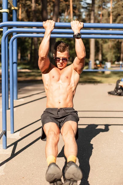 Sporty young male bodybuilder with muscular healthy body hanging on horizontal bar and doing exercise in the park