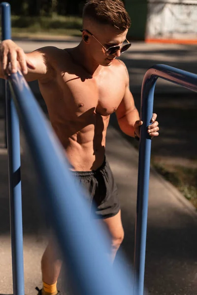 Cool sports athlete man with fitness muscular body with sunglasses and headphones listens to music and stands near the horizontal bars in the park