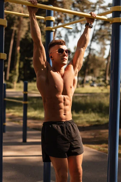 Strong sports man model with sunglasses and wireless headphones exercise and workout in the park. Athletic guy with muscular torso