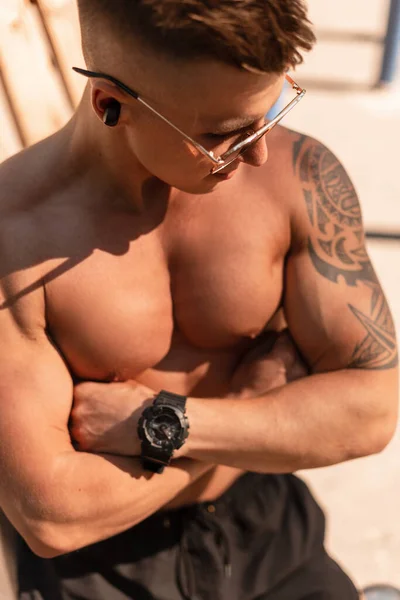 英俊强壮的男人 肌肉发达 戴着时髦的太阳镜 在沙滩上听着带有无线黑耳机的音乐 沐浴在阳光中 有纹身和运动手表的家伙 — 图库照片