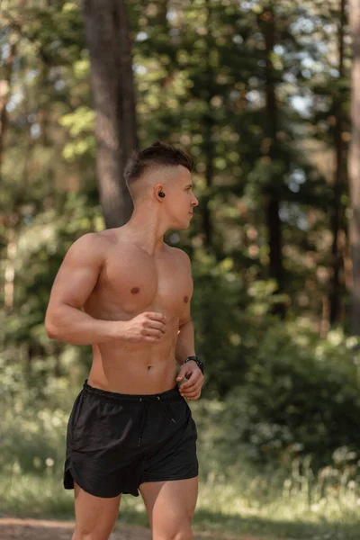 Athlete guy with muscular body runs in the park and listens to music in wireless headphones. Sport outdoors and healthy lifestyle