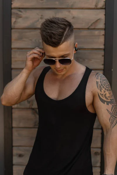 黒の流行のファッショナブルなTシャツの髪型とクールなファッションのヒップスター男性タトゥーは 木製の壁の近くにヴィンテージラウンドサングラススタンドを着用 — ストック写真