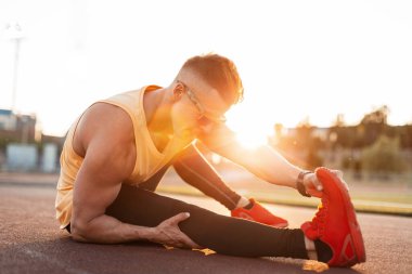 Güneş gözlüklü, saç stili ve spor ayakkabılı yakışıklı bir genç gün batımında stadyumda bacak esnetiyor.