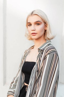 Şık yaz tişörtlü, kısa sarı saçlı ve siyah bluzlu moda kızı beyaz bir duvarın yanında duruyor.