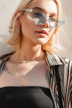 Mavi gözlüklü, gömlekli, siyah bluzlu güzel bir genç kızın güzel portresi ve güneş ışığında beyaz bir duvarın yanında.