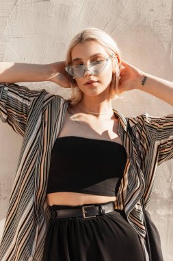 Moda gözlüklü, siyah bluzlu ve etekli güzel bir kadın model şehirde beyaz bir duvarın yanında poz veriyor.