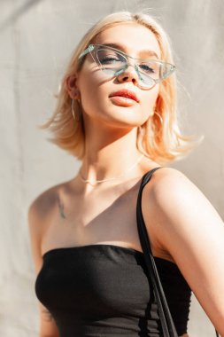 Güneşli bir yaz gününde, dışarıda gri bir duvarın yanında mavi güneş gözlüklü, göz kamaştırıcı genç sarışın kadın.