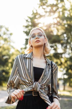 Yazlık gömlekli, siyah kolsuz tişörtlü, mavi gözlüklü güzel, mutlu genç kız parkta yürüyor.