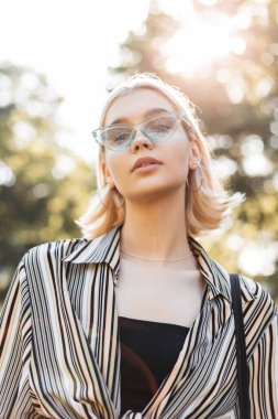 Şortlu, sarı saçlı, yaz kıyafetli, mavi güneş gözlüklü güzel bir kadın dışarıda yürüyor.