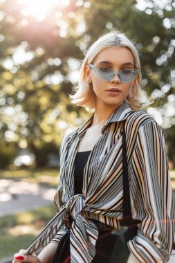 Şık, genç, sarışın, şık bahar kıyafetleri içinde, üzerinde gömlek, çanta ve gözlüklerle, güneşli bir günde parkta yürüyen şık bir kız.