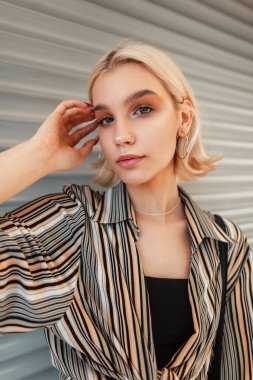 Moda çizgili tişörtlü, siyah bluzlu, kısa sarı saçlı güzel bir kızın şık bir portresi şehrin metal duvarının yanında duruyor ve kameraya bakıyor.