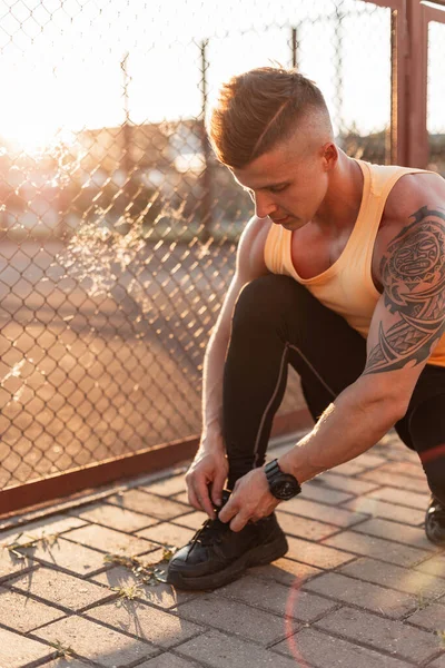 Fitness man tie shoelaces on the street at sunset. Athlete guy with a haircut and a tattoo getting ready for a workout
