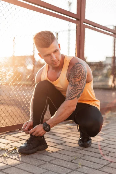 Fashionable handsome young man with hairstyle and tattoo on his shoulder tying his laces in sneakers on the street at sunset