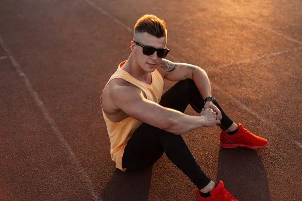 フィットネスハンサムな男運動選手でファッションサングラスでスポーツウェアで赤い靴座って 日没時にトレッドミルで休む — ストック写真