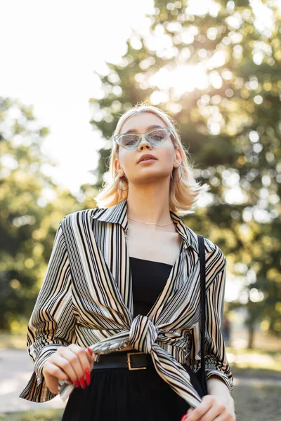 Yazlık gömlekli, siyah kolsuz tişörtlü, mavi gözlüklü güzel, mutlu genç kız parkta yürüyor.