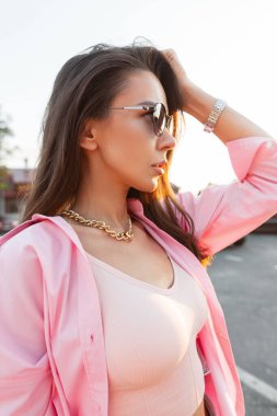 Günbatımında şehirde güneş gözlüklü, şık, pembe elbiseli, alımlı, alımlı, genç ve çekici bir kadın. Bahar günü açık havada günlük kıyafetleriyle modaya uyan beyaz kız.