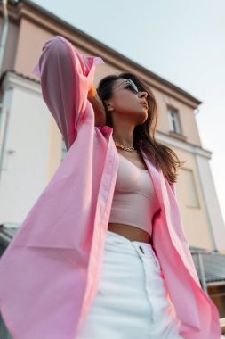 Havalı güneş gözlüklü, pembe gömlekli, üst ve beyaz kotlu göz kamaştırıcı genç bir kadın.