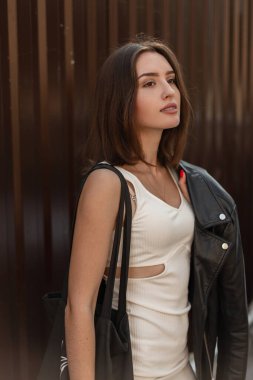 Havalı beyaz elbiseli, deri ceketli, siyah çantalı, metal duvarın yanında yürüyen şık, genç ve güzel bir hippi kız.