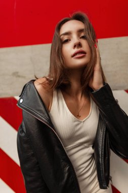 Kısa saçlı, hoş yüzlü, modaya uygun beyaz elbiseli, siyah deri ceketli seksi bir genç kadın kırmızı ve beyaz duvarın yanındaki kameraya bakıyor.