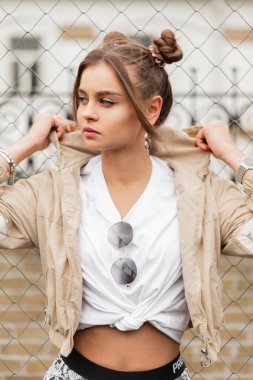 Modaya uygun sokak spor kıyafetleri içinde, rüzgarlık ve beyaz tişörtlü güzel bir kız sokakta metal şebekenin yanında duruyor.