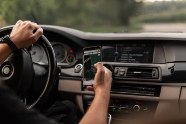 Genç adam araba kullanıyor ve elinde harita navigasyon uygulaması olan bir akıllı telefon tutuyor.